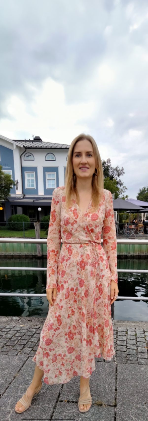 Liubov, 38 Jahre alt, steht in einem langen, geblümten Kleid an einem Gewässer mit einem Gebäude im Hintergrund. Ukrainische Single Frauen, die eine ernsthafte Beziehung suchen.