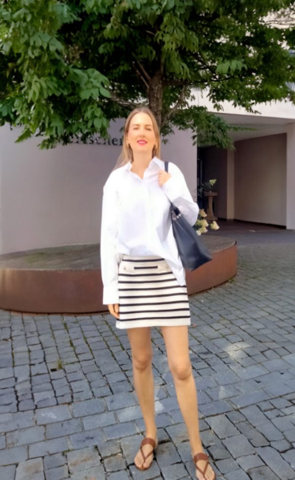 Liubov, 38 Jahre alt, lächelt in die Kamera. Sie trägt eine weiße Bluse, einen gestreiften Rock und eine schwarze Handtasche und steht auf einem gepflasterten Platz mit einem Baum. Ukrainische Single Frauen, die eine ernsthafte Beziehung suchen.