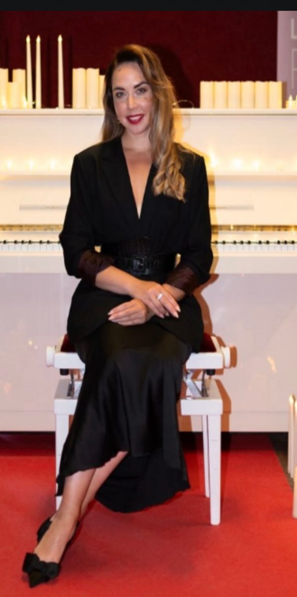 Antonina, 38 Jahre alt, sitzt elegant auf einem Klavierhocker in einem schwarzen Outfit, vor einem weißen Klavier mit Kerzen. Diese stilvolle Dame aus Osteuropa sucht über eine Partnervermittlung ihren passenden Partner.